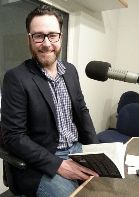 Craig A. Hart holding a book beside a broadcast microphone during a live radio reading